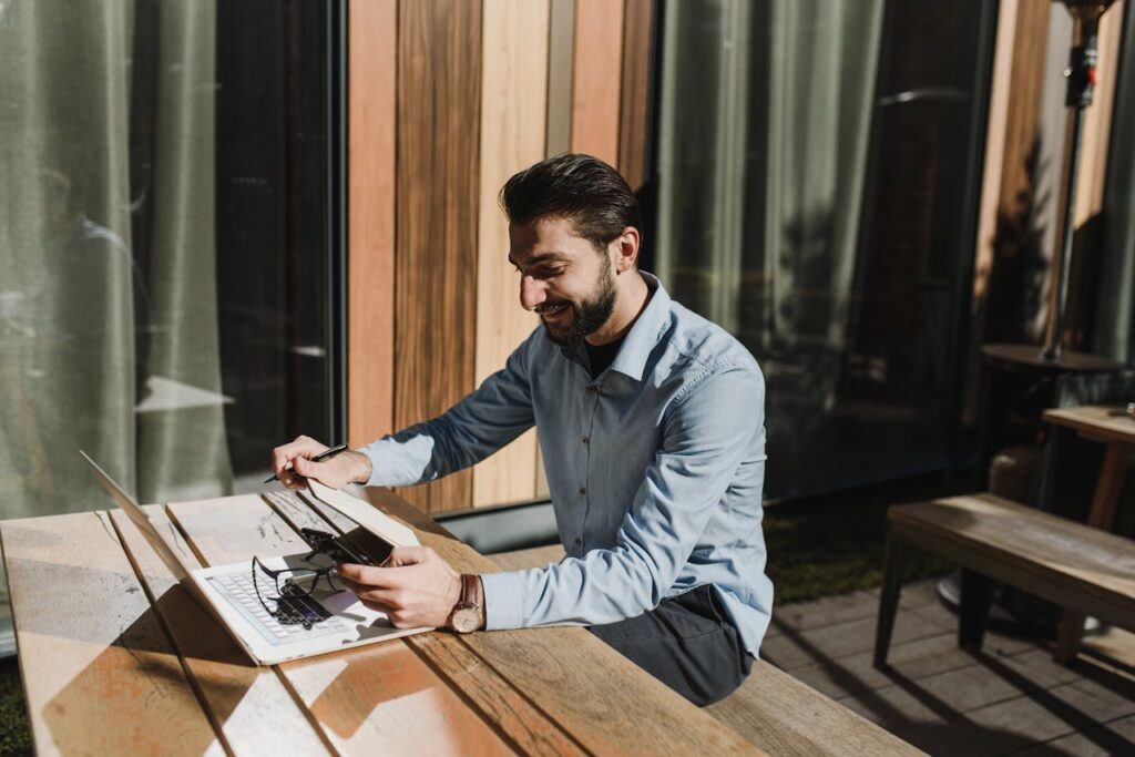 Bearded man using laptop and smartphone outdoors as a remote freelancer during the day.